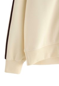 Krémová mikina s žebrovanými manžetami, hladkým povrchem a kontrastním tmavým pruhem po straně pro zvýraznění detailu.