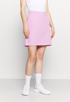 Kobieta ubrana w różową spódnicę do kolan, biały top, białe skarpetki i białe buty Nike, stojąca na tle gładkiej białej ściany.