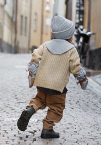 Beige gestrickter Pullover mit grauem Kragen, braune Cordhose, graue Strickmütze und schwarze Stiefel. Kind hält eine gemusterte Tasche und läuft auf Kopfsteinpflaster.