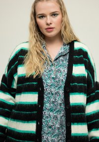 Schwarz und türkis gestreifter Cardigan mit weicher Textur, mit Knöpfen und oversized Passform, über einem mit Paisleymuster bedruckten Hemd getragen.