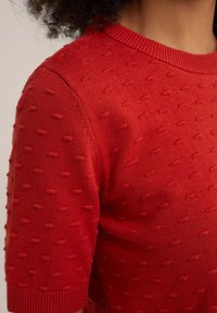 Close-up of a red textured knit short sleeve sweater worn by a person with curly hair, showing shoulder and upper chest details.