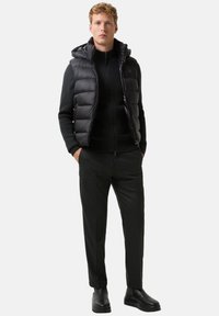 Schwarze gesteppte Weste mit Kapuze über einem schwarzen Reißverschluss-Pullover, kombiniert mit dunkler Hose und schwarzen Stiefeln. Glatte Textur, minimalistisches Design.