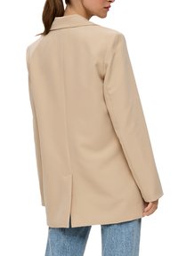 Beige Blazer mit strukturiertem Kragen, taillierter Passform und Schlitz auf der Rückseite, kombiniert mit hellblauen Jeans. Glattes, leichtes Material.
