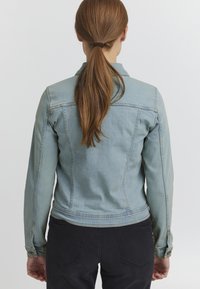 Ljusblå denimjacka med strukturerade sömmar, knäppta manschetter och en figursydd siluett. Bakifrån syns kragen och rak nederkant.