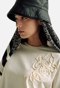 Černý klobouk s kbelíkovým tvarem s detailem šátku s vzorem, doplněný krémově zbarveným topem s vyšívaným květinovým motivem z texturovaných nití.