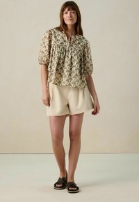 Korte mouwen, beige blouse met een bladerenpatroon; gecombineerd met lichte shorts en donkere sandalen. Glad materiaal met een ontspannen pasvorm.