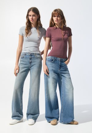 Due modelli indossano top aderenti a maniche corte, uno grigio e uno borgogna, abbinati a jeans a vita alta e gamba larga di colore blu chiaro e scarpe casual.