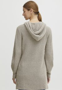 Beige gestrickter Hoodie mit geripptem Muster, ausgestattet mit einer großen, lässig fallenden Kapuze und langen Ärmeln. Das Kleidungsstück wirkt weich und warm.
