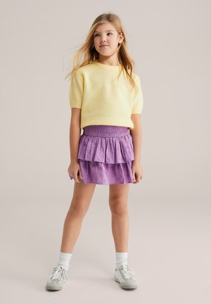 Gul strikket kortærmet sweater kombineret med en lilla flerlags nederdel med flæser, der har et polkaprikket mønster, samt grå sneakers med hvide sokker.