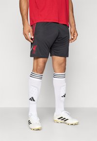 Shorts de sport noirs avec le logo rouge de Liverpool FC, associés à des protège-tibias blancs avec des rayures noires, et des chaussures de football blanches avec des accents noirs.