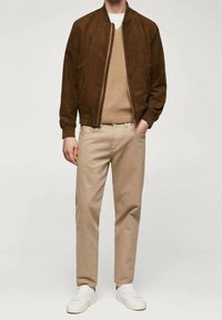 Chaqueta bomber de ante marrón sobre un suéter de punto beige, combinada con pantalones beige claros y zapatillas blancas, creando un look casual en capas.