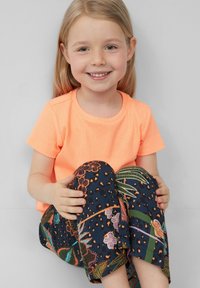 Neon oranje geribde T-shirt met korte mouwen, gecombineerd met marineblauwe broek met een patroon van bloemen en bladeren. Kind dat zit en lacht.