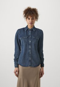 Mørkeblå denimskjorte med naturlig krage, knappelukking, to brystlommer og lange ermer. Stoffet har en myk, noe teksturert finish.