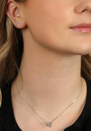 Silberne herzförmige Ohrstecker mit klaren Steinakzenten an einem Ohr und einer passenden zarten Halskette mit Herzanhänger.