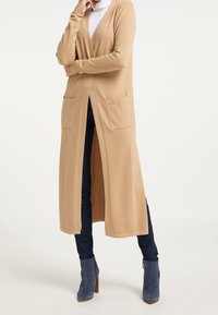 Cardigan long beige en matière douce, avec une ouverture frontale profonde, deux poches avant et des manches longues. Associé à un jean foncé et des bottines bleues.
