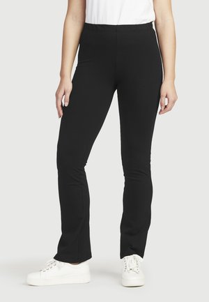 Schwarze Stretch-Leggings mit glatter Textur, im figurbetonten Design und elastischem Taillenbund, kombiniert mit weißen Sneakers.