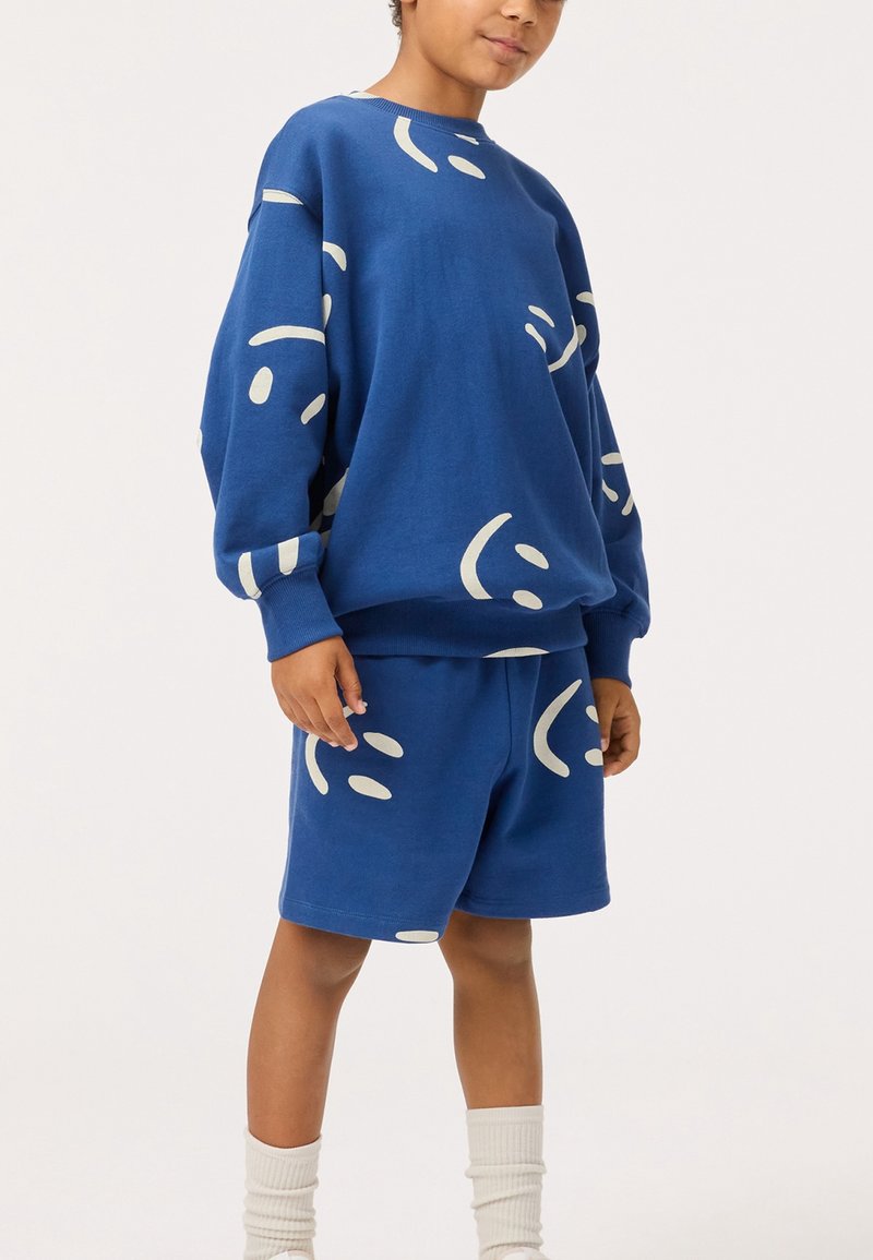 Enfant portant un sweat-shirt bleu et un short avec des motifs abstraits de visages souriants blancs, assortis de chaussettes blanches, debout devant un fond uni.