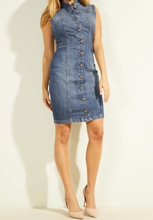 Femme portant une robe en jean sans manches boutonnée jusqu'aux genoux et des talons hauts beiges, debout devant un fond clair uni.