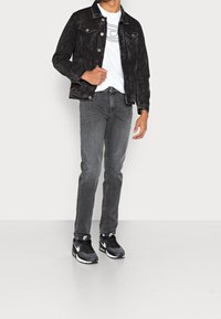 Chaqueta de mezclilla negra con dos bolsillos frontales, camiseta gráfica blanca, pantalones grises y zapatillas Nike negras con detalles blancos.