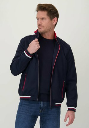 Marine bomberjack met rode accenten, ritssluiting, ribgebreide boorden met witte en rode strepen, en zijzakken, gedragen over een donkere top.