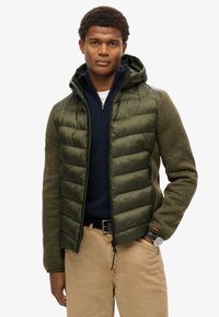 Giacca imbottita verde oliva con cappuccio, maniche strutturate e tasche con zip; indossata sopra un pullover blu navy e pantaloni beige.