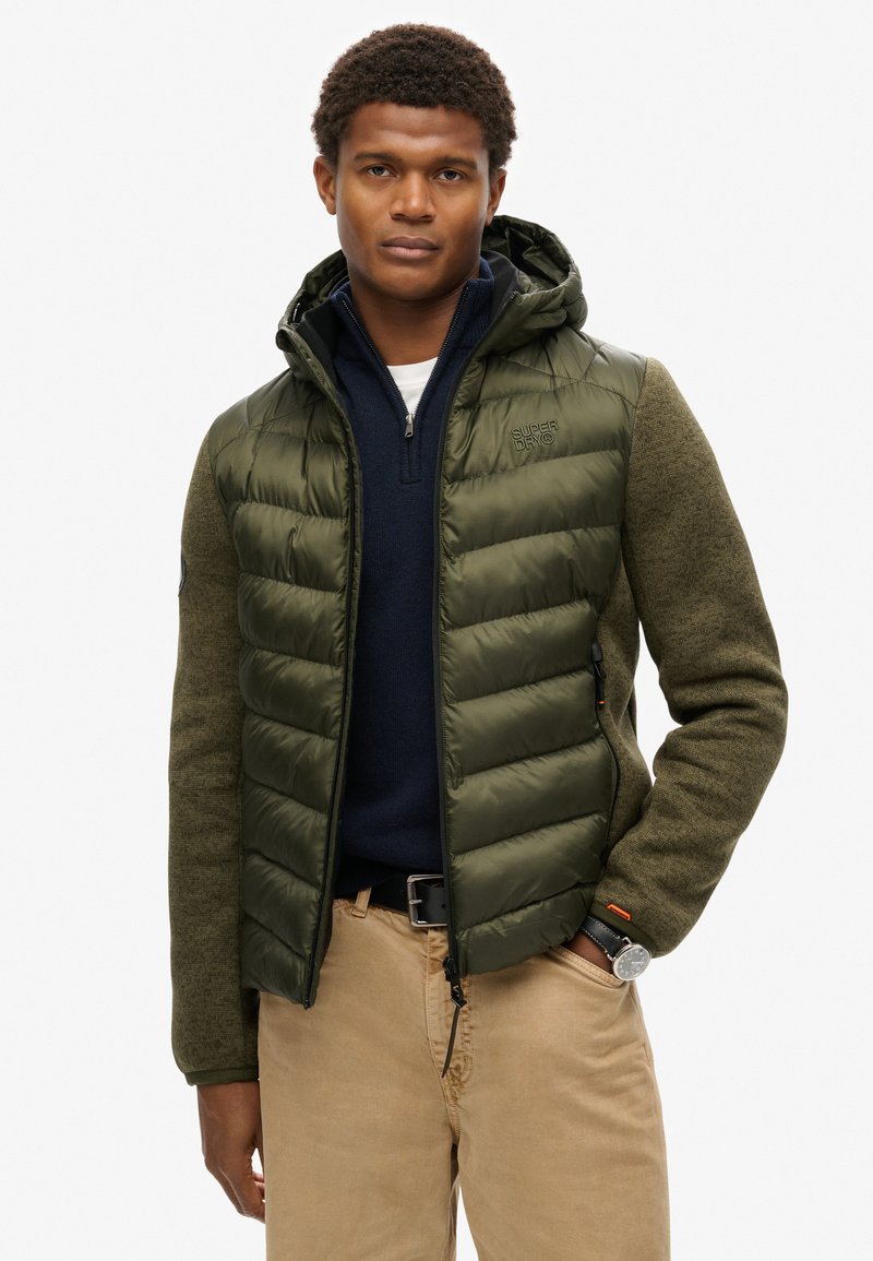Giacca imbottita verde oliva con cappuccio, maniche strutturate e tasche con zip; indossata sopra un pullover blu navy e pantaloni beige.