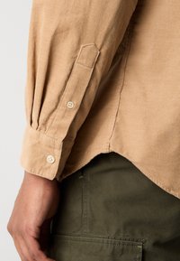 Braune Cordhemd mit langen, gepufften Ärmeln und geknöpften Manschetten. Der Saum ist leicht geschwungen. Über einem olivgrünen Stoff getragen.