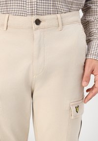 Beżowe spodnie cargo z zapięciem na guzik, bocznymi kieszeniami i żółtym haftowanym logo na lewej kieszeni. Trwała bawełniana tkanina, gładka tekstura.