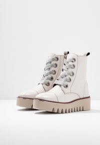 Valkoiset patenttinahkaiset nilkkurit harmailla nauhoilla, tasapohjalla ja siksak-kuviolla. Varustettu metallisilla nysteillä ja vetolenkillä.