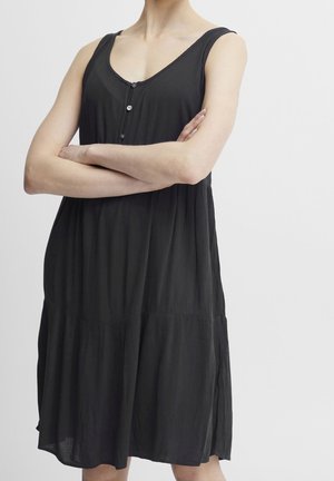 Mujer con vestido negro sin mangas con botones en el frente, brazos cruzados, sobre fondo blanco liso.