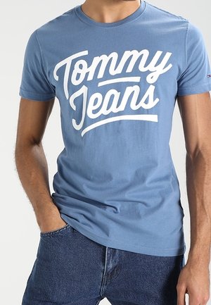 Blauwe katoenen T-shirt met vetgedrukte witte tekst "Tommy Jeans" in een cursieve lettertype. Korte mouwen en een ronde hals. Draagt het in combinatie met een spijkerbroek.