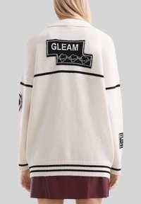 Kobieta w białym, oversizowym swetrze w czarne paski z naszywkami "GLEAM", w połączeniu z bordową spódnicą, widoczna z tyłu.