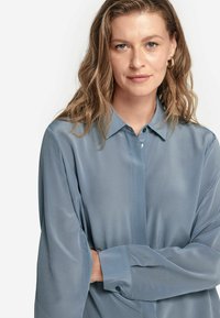Blusa azzurra chiara con maniche lunghe, realizzata in un tessuto morbido, con colletto e polsini abbottonati. Presenta un design pulito e minimalista.