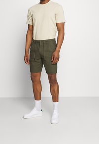 T-shirt beige a maniche corte con piccolo logo, abbinato a pantaloni corti verde oliva con vita regolabile. Sneakers bianche e calze completano l'outfit.