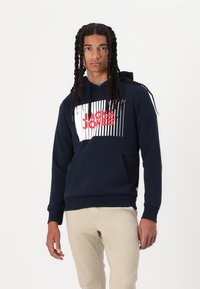 Felpa blu navy in tessuto spesso con un motivo grafico bianco e rosso, che presenta il testo "JACK & JONES" e linee verticali. Tasca a marsupio frontale.