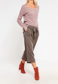 Gruba dzianinowa sweter w kolorze różowym, szerokie spodnie oliwkowe z wiązaniem z przodu oraz czerwone botki za kostkę. Teksturowany materiał, casualowy design, luźny krój.