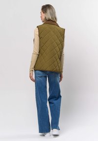 Quiltet olivengrønn vest med brun krage, brukt over en beige langermet topp, kombinert med lys blå vide jeans og hvite joggesko.