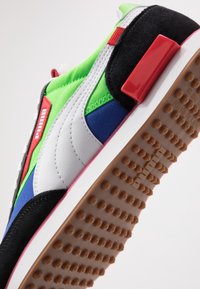 Baskets de sport présentant un mélange de matériaux textiles verts, bleus et noirs, avec un accent en cuir blanc et des éléments rouges. Semelle marron texturée.