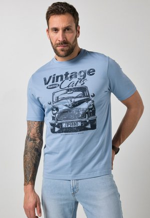Mann mit tätowiertem Arm trägt hellblaues T-Shirt mit Vintage-Auto-Grafik und dem Text „Vintage Racing Cars“ sowie hellblaue Jeans, steht vor einfachem Hintergrund.