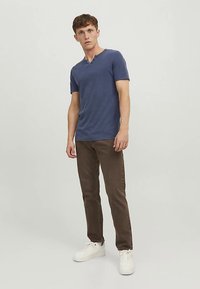 Tumši zils T-krekls ar V veida izgriezumu, izgatavots no mīksta auduma, kombinēts ar brūnām džinsām un baltām sporta kurpēm. Modelis stāv taisni priekšā gaišā fona.