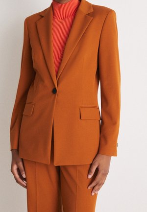 Frau trägt einen eng anliegenden, verbrannt-orangefarbenen Blazer mit Kissing Lapels und einem schwarzen Knopf über einem roten, gerippten Rollkragenpullover.