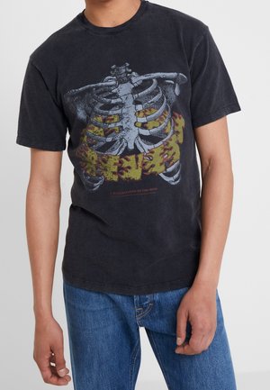 Personne portant un t-shirt noir avec un motif de cage thoracique et des flammes jaunes, associé à un jean bleu sur un fond uni.