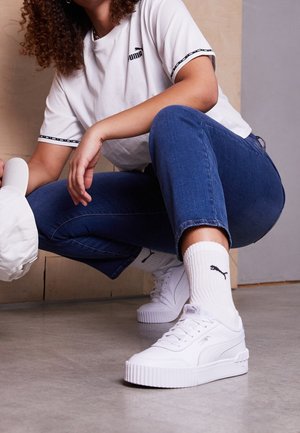 Bílé platformové tenisky s texturovanou podrážkou, spárované s bílými pruhovanými ponožkami s černým logem. Model má na sobě bílé tričko s černým lemem a modré džíny.