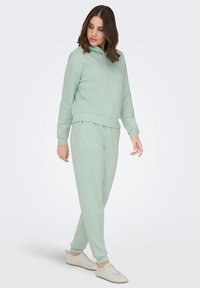Svetlo zelena jopa s kapuco in žepom kengurujčkom, združena z ujemajočimi se trenirkami, oba izdelana iz mehke tkanine, z manšetami in nastavljivo kapuco.