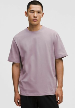 Giovane uomo con capelli corti che indossa una maglietta oversize di colore lilla chiaro e pantaloni neri, in piedi davanti a uno sfondo neutro.