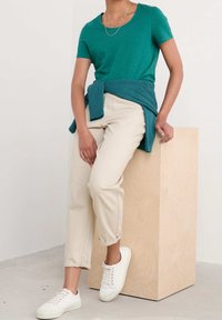 Tyrkysové tričko s krátkým rukávem v kombinaci s pleteným kardigánem, krémové zúžené kalhoty a bílé tenisky; pózující na světle dřevěném bloku.