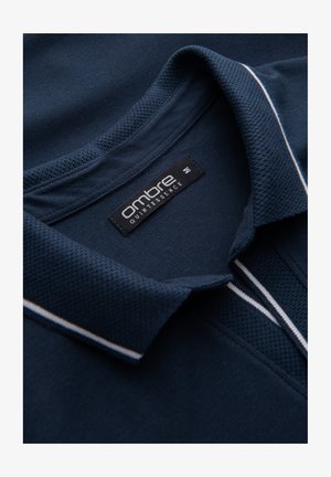 Laivastonsininen polo-paita, jossa on rakenteellinen kangas, kauluksellinen kaula ja valkoiset korostusraidat kauluksessa. Tunnuksessa lukee "ombre quintessence."