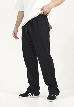 Pantalons de survêtement noirs avec une taille élastique et des cordons ajustables. Tissu doux, coupe décontractée, associés à des baskets noires et une chemise blanche.