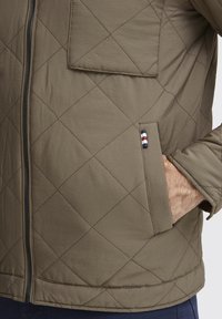Quilted olive green jacket with diagonal stitching, zip closure, and a side pocket featuring striped accent detail. Soft, textured fabric.