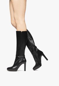 Bottes noires en cuir montantes avec une finition brillante, un talon aigu et un accent de boucle. Elles présentent un design épuré avec une texture lisse.